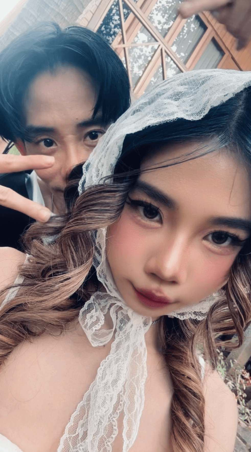 Her in a lace veil and bridal makeup, him behind her with a peace sign